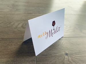 A white folded notecard stands on a gray wood floor. The card reads, "merry Merlot" in a script font and is accented with a wine glass that has a snowflake inside it. Christmas Cheers by Stationare.