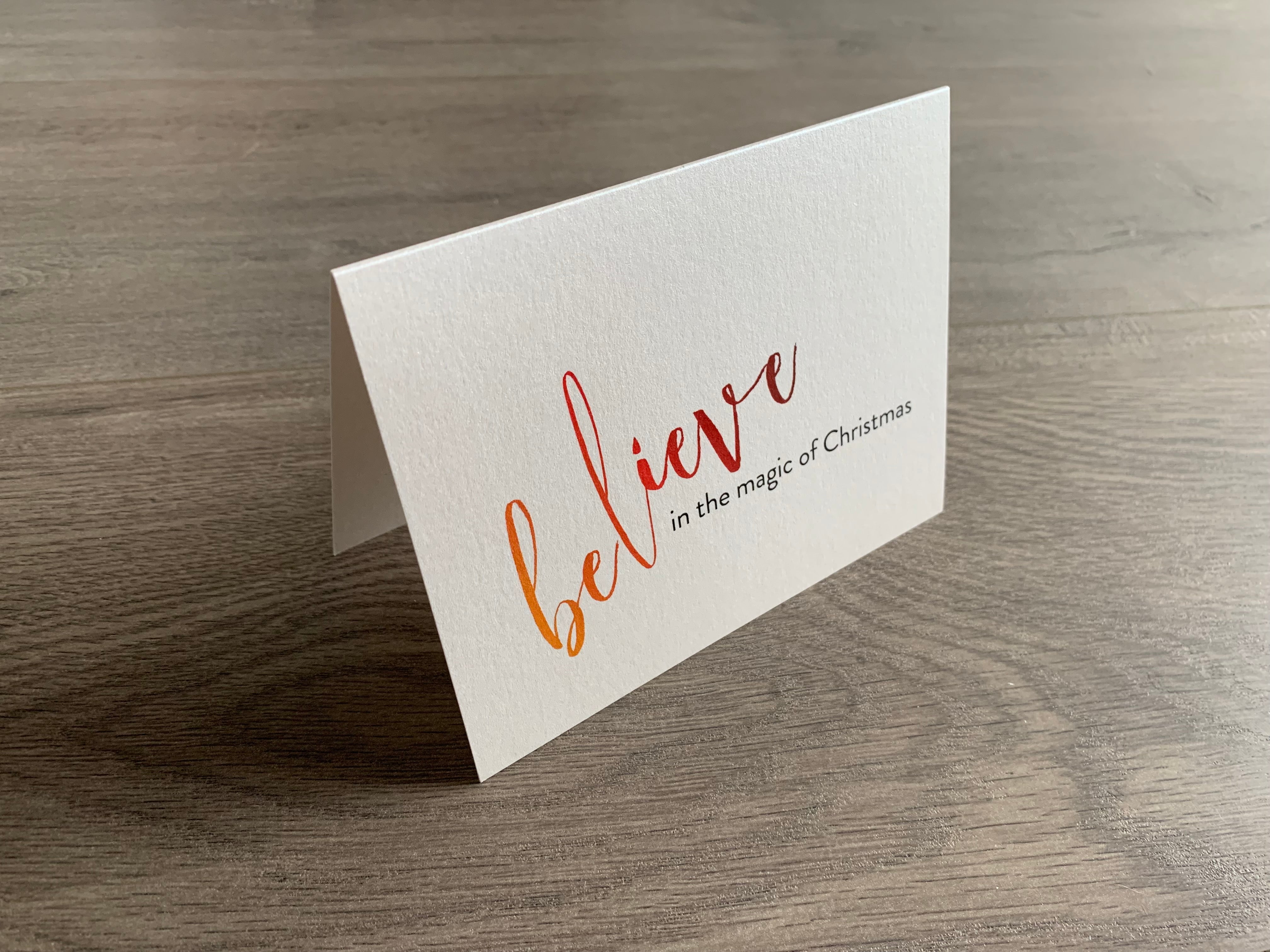 A folded notecard is propped up on a gray wood floor. The notecard is of a cream-colored pearlized paper. The card says, "Believe in the magic of Christmas." Meaning of Christmas collection by Stationare.