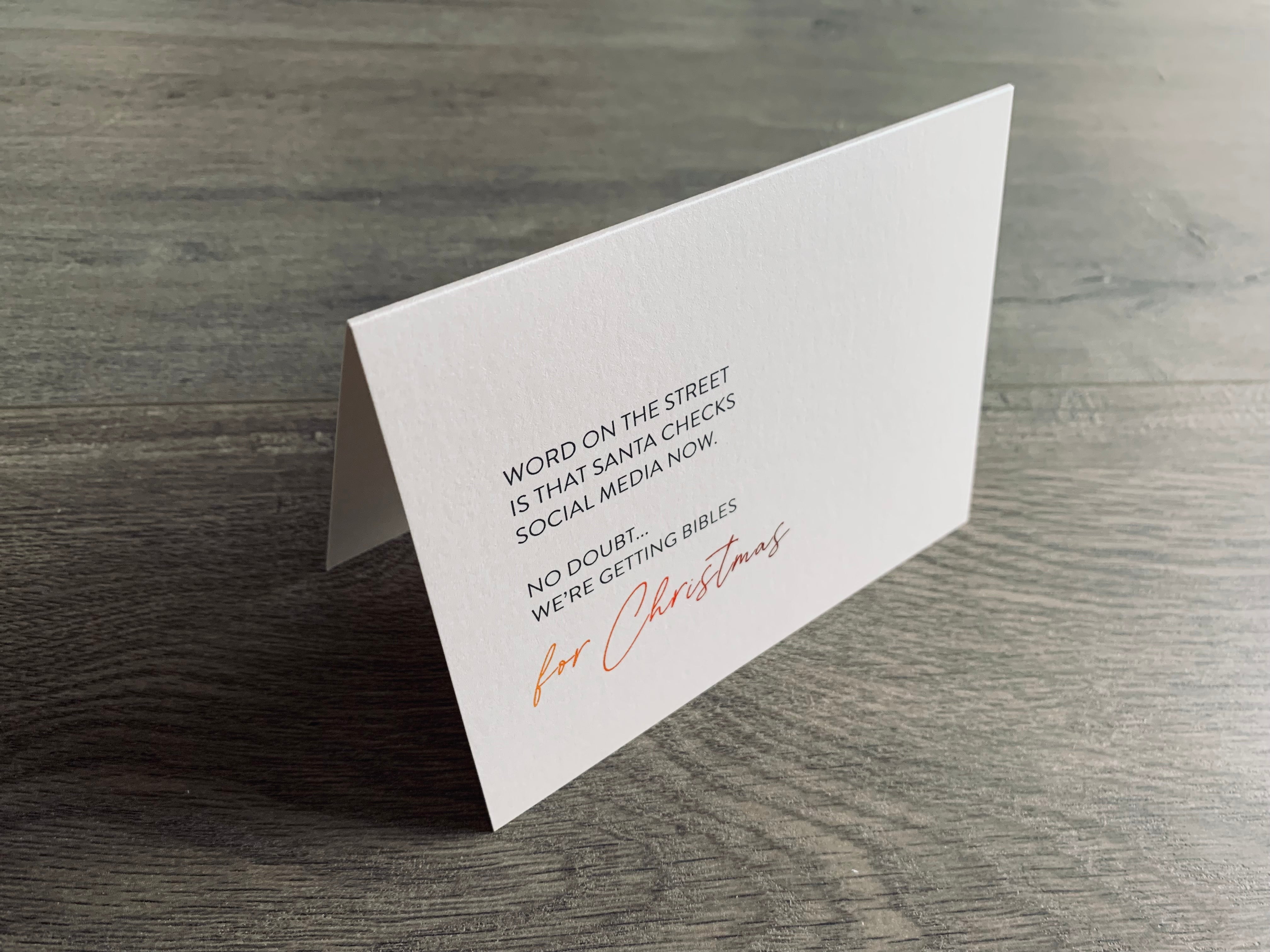 A folded notecard is propped up on a gray wood floor. The notecard is of a cream-colored pearlized paper. The card says, "Word on the street is that Santa checks social media now. No doubt... we're getting bibles for Christmas." Christmas Chuckles collection by Stationare.