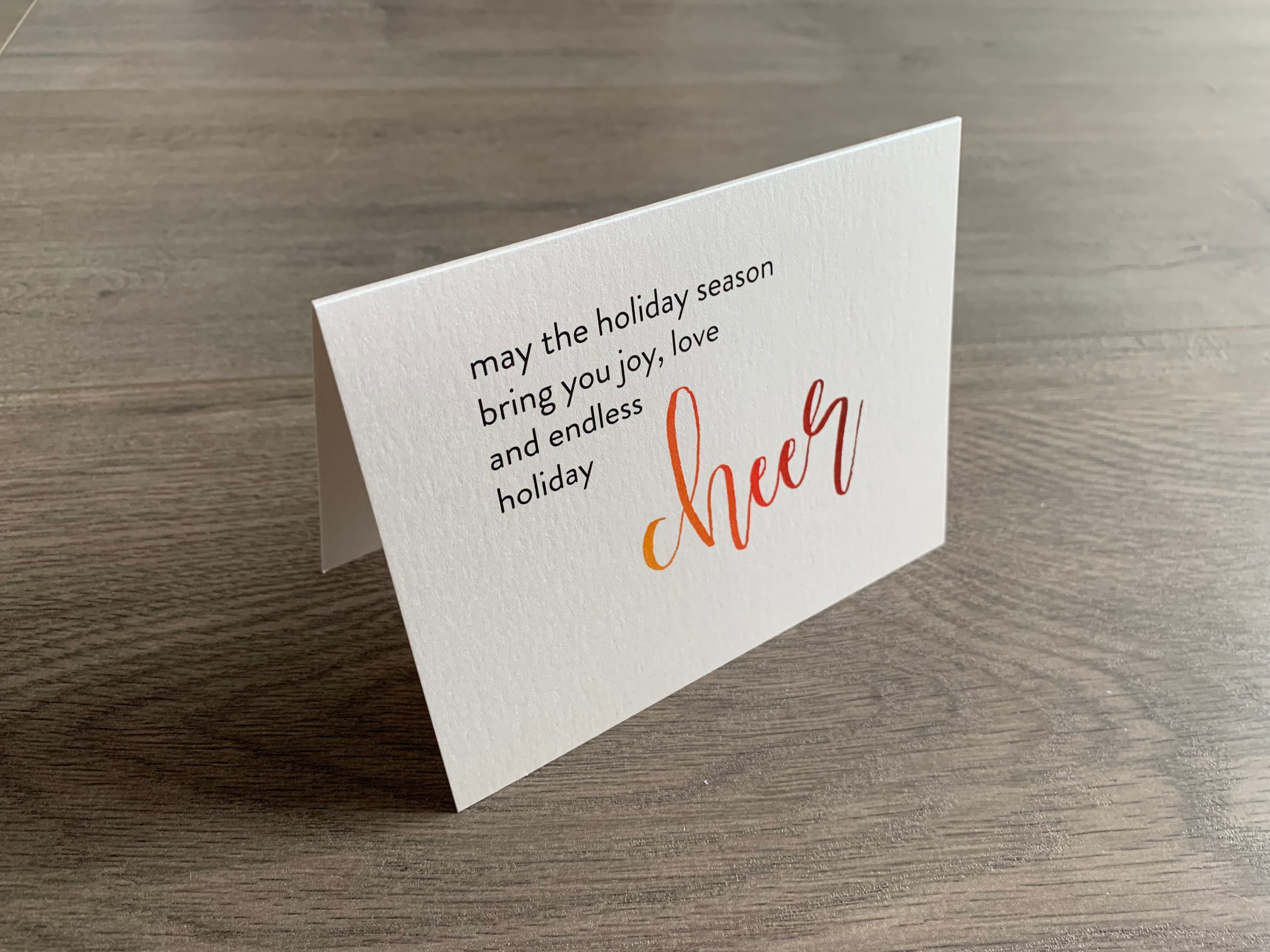 A folded notecard is propped up on a gray wood floor. The notecard is of a cream-colored pearlized paper. The card says, "may the holiday season bring you joy, love and endless holiday cheer" Meaning of Christmas collection by Stationare.