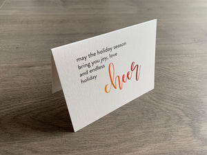 A shimmery cream notecard is propped up on a wooden floor. The card says, "may the holiday season bring you joy, love and endless holiday cheer" The word "cheer" is in a script font that fades from orange to red. Meaning of Christmas collection by Stationare.