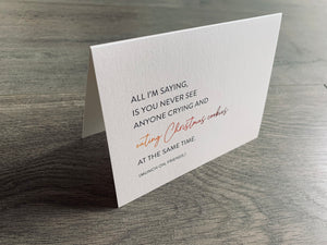 A folded notecard is propped up on a gray wood floor. The notecard is of a cream-colored pearlized paper. The card says, "All I'm saying, is you never see anyone crying and eating Christmas cookies at the same time. Munch on, friends." Christmas Chuckles collection by Stationare.