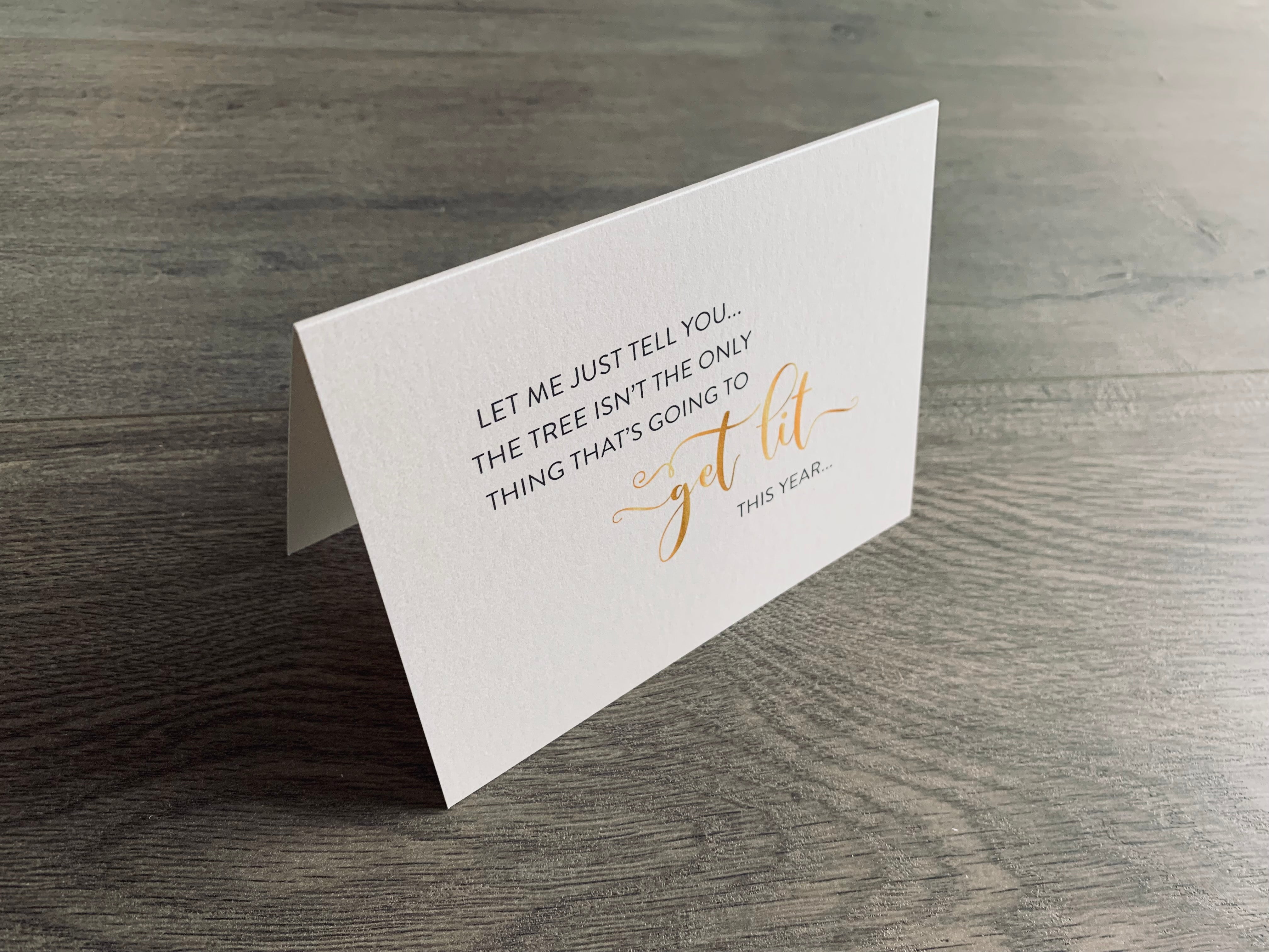 A folded notecard is propped up on a gray wood floor. The notecard is of a cream-colored pearlized paper. The card says, "Let me just tell you... the tree isn't the only thing that's going to get lit this year." Christmas Chuckles collection by Stationare.