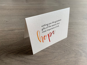 A shimmery cream notecard is propped up on a wooden floor. The card says, "wishing you the greatest gifts of the season but most important of all, hope." The word "hope" is in a script font that fades from orange to red. Meaning of Christmas collection by Stationare.