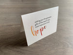 A folded notecard is propped up on a gray wood floor. The notecard is of a cream-colored pearlized paper. The card says, "wishing you the greatest gifts of the season but most important of all, hope" Meaning of Christmas collection by Stationare.