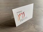 A folded notecard is propped up on a gray wood floor. The notecard is of a cream-colored pearlized paper. The card says, "wishing you joy and cheer now and through the new year" Meaning of Christmas collection by Stationare.