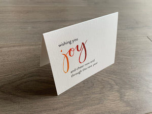 A shimmery cream notecard is propped up on a wooden floor. The card says, "wishing you joy and cheer now and through the new year." The word "joy" is in a script font that fades from orange to red. Meaning of Christmas collection by Stationare.