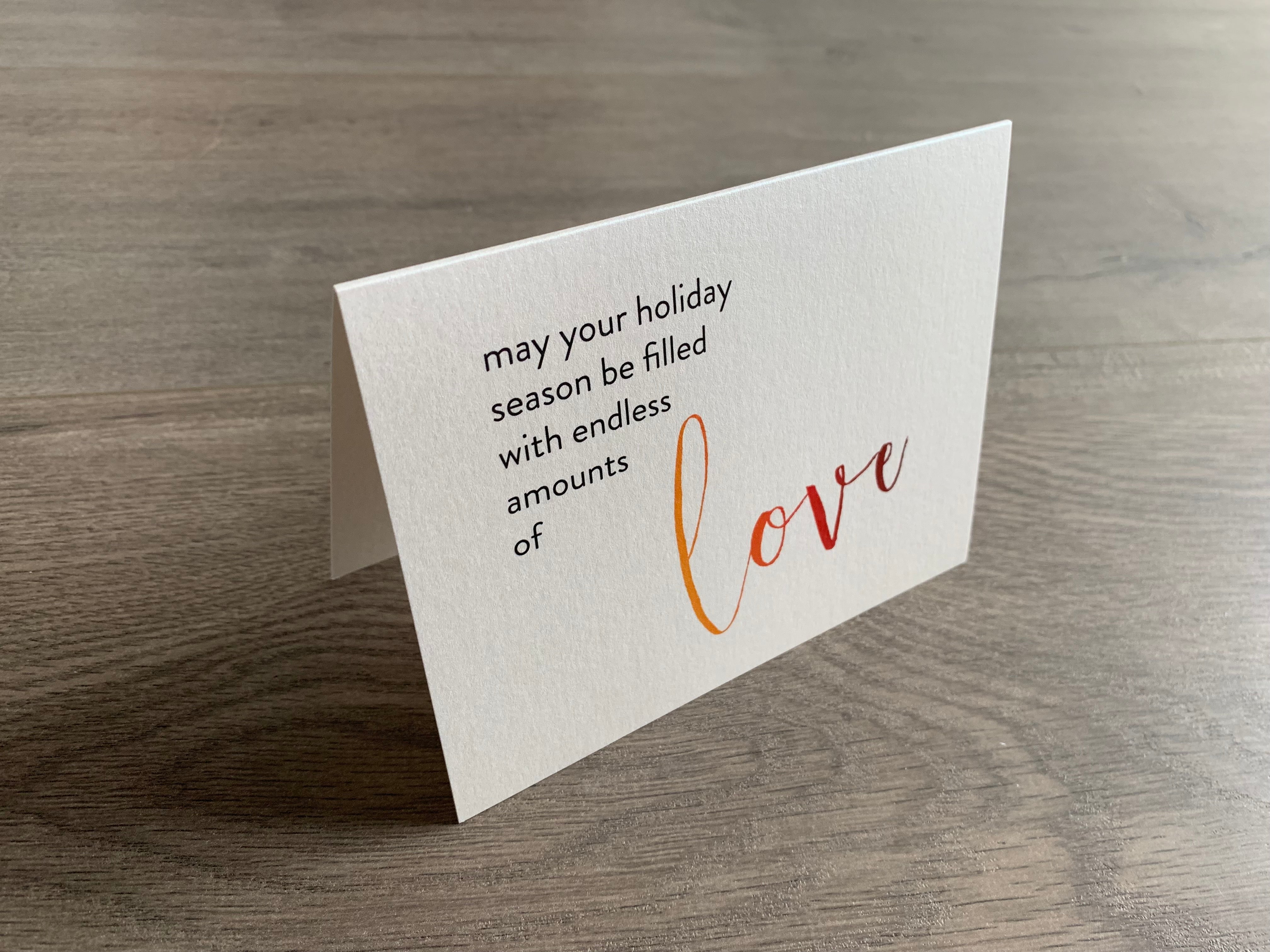 A shimmery cream notecard is propped up on a wooden floor. The card says, "may your holiday season be filled with endless amounts of love." The word "love" is in a script font that fades from orange to red. Meaning of Christmas collection by Stationare.