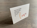 A shimmery cream notecard is propped up on a wooden floor. The card says, "may your holiday season be filled with endless amounts of love." The word "love" is in a script font that fades from orange to red. Meaning of Christmas collection by Stationare.