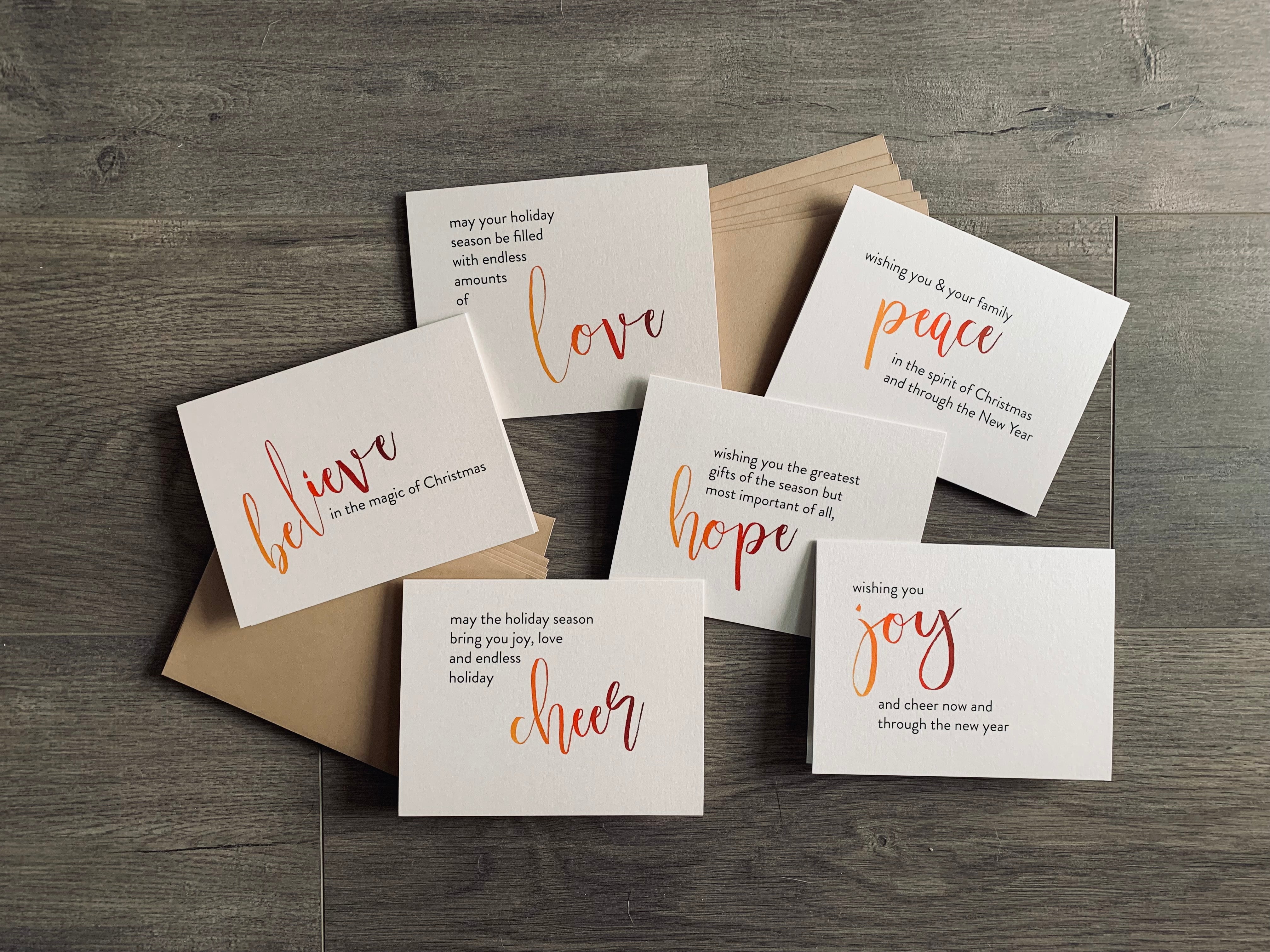 The 6 cards from the Meaning of Christmas collection lie on a gray wooden background with two stacks of kraft envelopes. The cards are printed on a cream pearlized paper. Each card has a large script word in an ombre of orange to burgundy. The rest of the sentiment is in black sans serif font.