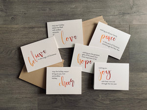 The 6 cards from the Meaning of Christmas collection lie on a gray wooden background with two stacks of kraft envelopes. The cards are printed on a cream pearlized paper. Each card has a large script word in an ombre of orange to burgundy. The rest of the sentiment is in black sans serif font.