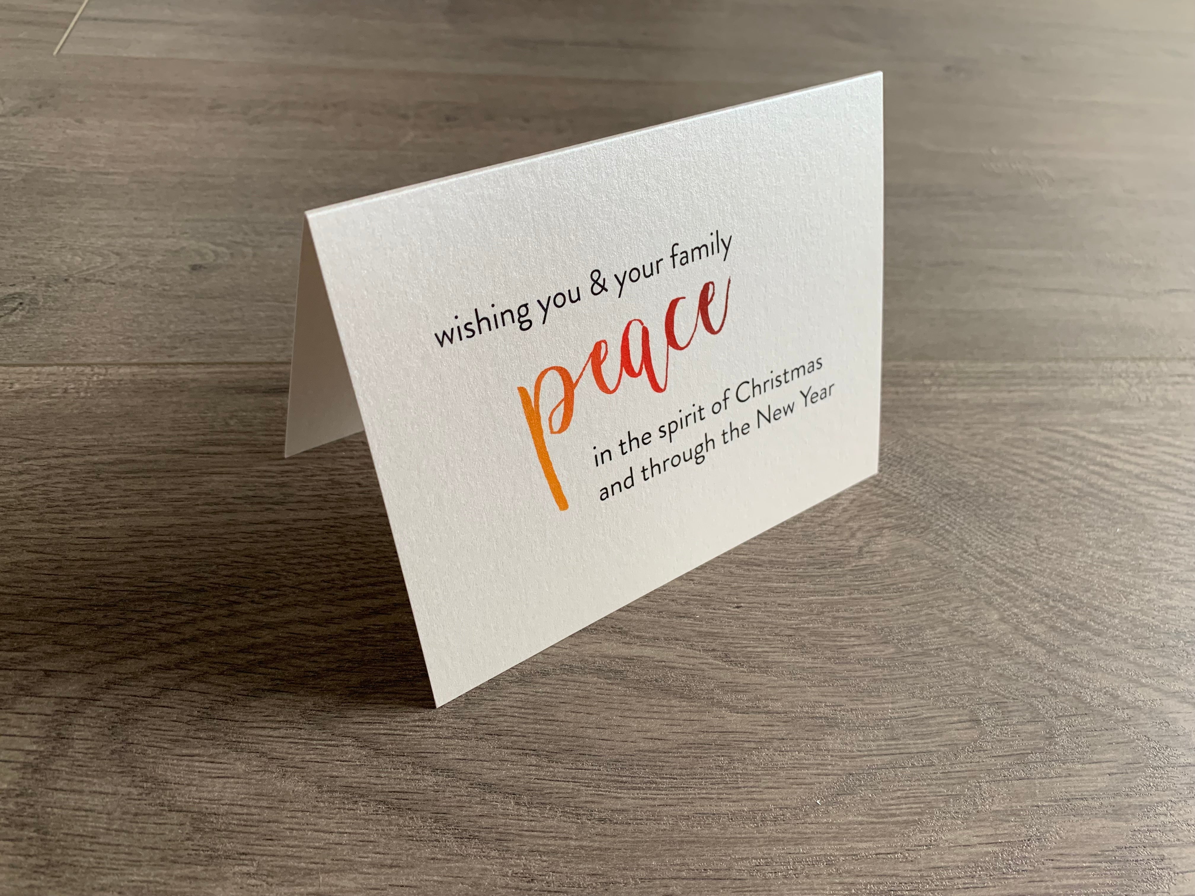 A shimmery cream notecard is propped up on a wooden floor. The card says, "wishing you and your family peace in the spirit of Christmas and through the new year." The word "peace" is in a script font that fades from orange to red. Meaning of Christmas collection by Stationare.
