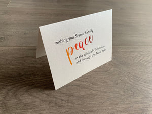 A folded notecard is propped up on a gray wood floor. The notecard is of a cream-colored pearlized paper. The card says, "wishing you and your family peace in the spirit of Christmas and through the New Year." Meaning of Christmas collection by Stationare.