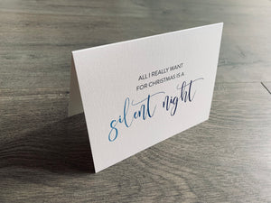 A folded notecard is propped up on a gray wood floor. The notecard is of a cream-colored pearlized paper. The card says, "All I really want for Christmas is a silent night" Christmas Chuckles collection by Stationare.