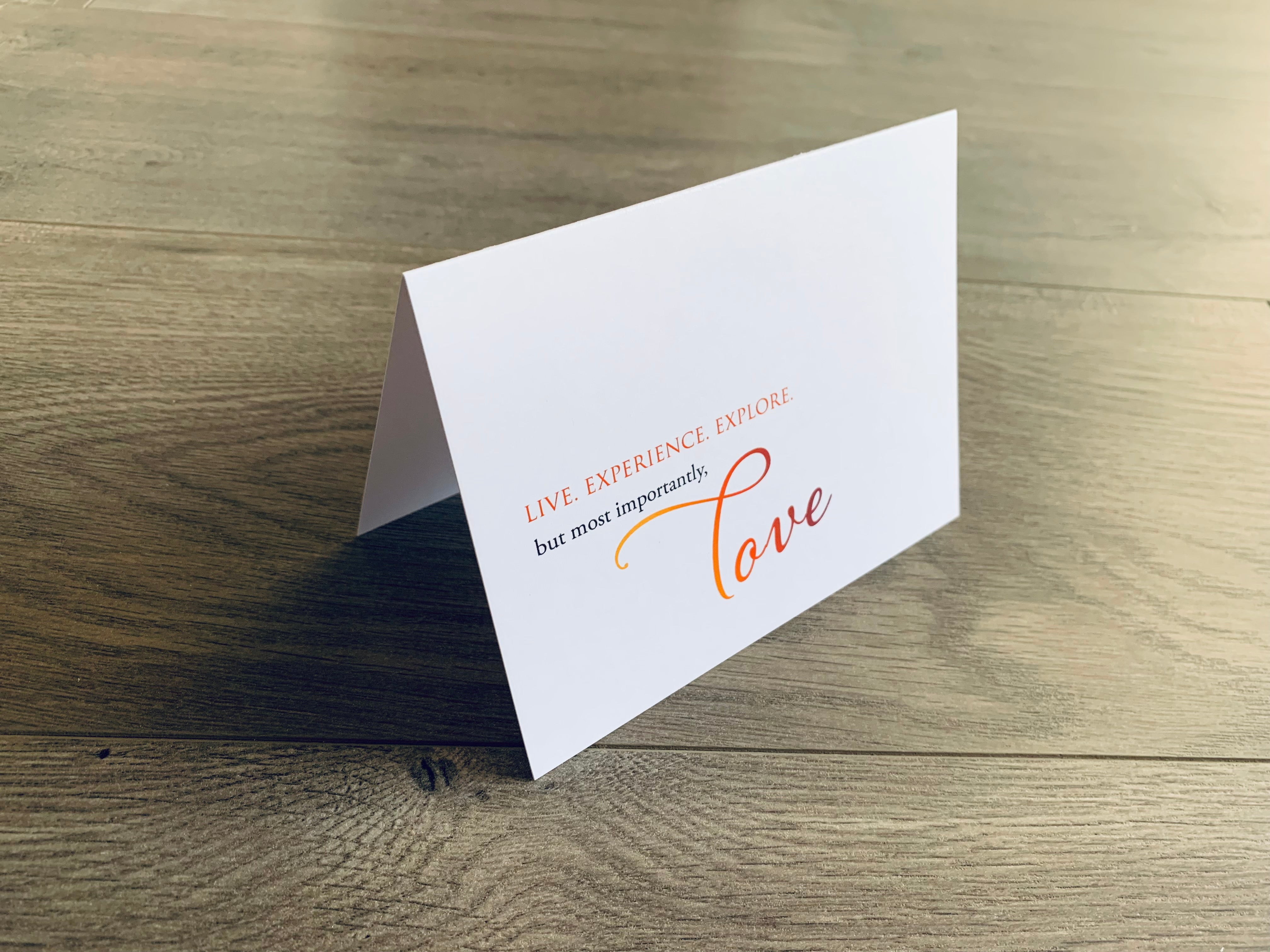 A folded white notecard is propped up on a wooden floor. The card reads, "live. experience. explore. but most importantly, love" in a mix of serif and script fonts. Love Collection by Stationare.