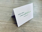 A white folded notecard is propped up on a wooden floor. The card has the Irish proverb, "many a time... a man's mouth broke his nose." From the Irish Laughs Collection by Stationare.