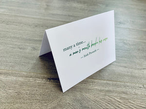 A white folded notecard is propped up on a wooden floor. The card has the Irish proverb, "many a time... a man's mouth broke his nose." From the Irish Laughs Collection by Stationare.
