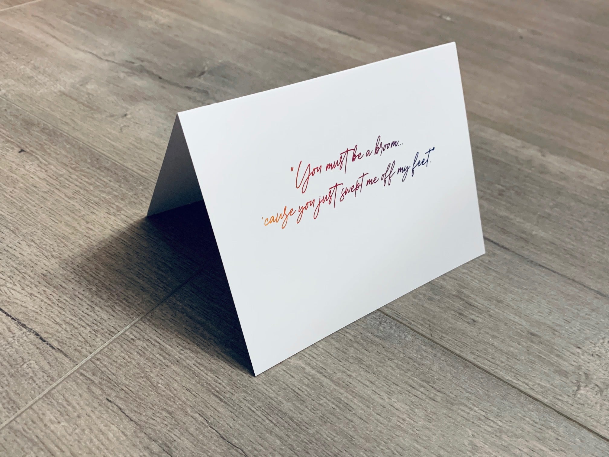 A folded white notecard is propped up on a wooden floor. The card reads, "You must be a broom... 'cause you just swept me off my feet." Stationare's Pick Up Lines collection.