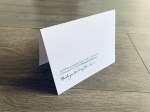 A folded white notecard is propped up on a wooden floor. The card reads, "During challenging times, I realize how much I need friends like you. Thank you for being there for me." Stationare's Surviving Hard Times collection.