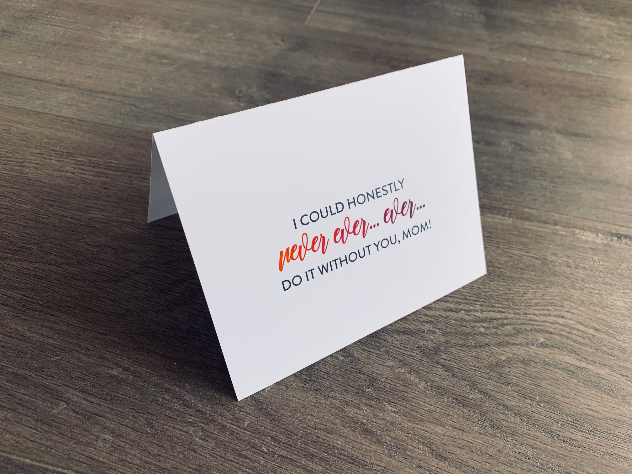 A white folded notecard is propped up on a gray wooden floor. The card says, "I could honestly never ever... ever... do it without you, Mom!" Mother's Day card by Stationare.