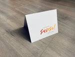 A white card is propped on a gray wood floor. The card reads, "I never met a sunset I didn't like..." Beach Life Collection by Stationare.
