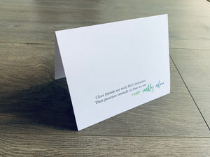 A white, folded notecard sits on a wooden floor. The card says, "Close friends are truly life's treasures. Their presence reminds us that we are never really alone." The Friendship collection by Stationare.