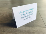 A white, folded notecard sits on a gray wooden floor. The card reads, "When you have a hard time finding the bright side, I will gladly sit next to you in the dark." Sending Support collection by Stationare.