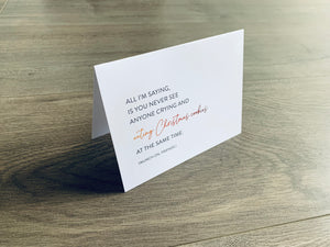 A white folded notecard is propped up on a wooden floor. On the front of the card, it says "All I'm saying, is you never see anyone crying and eating Christmas cookies at the same time. Munch on, friends." Christmas Chuckles collection by Stationare.