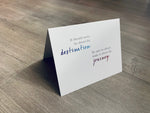 A folded white notecard is propped up on a wooden floor. The card reads, "It should never be about the destination. Be sure to always make it about the journey." Stationare's Travel Lovers collection.