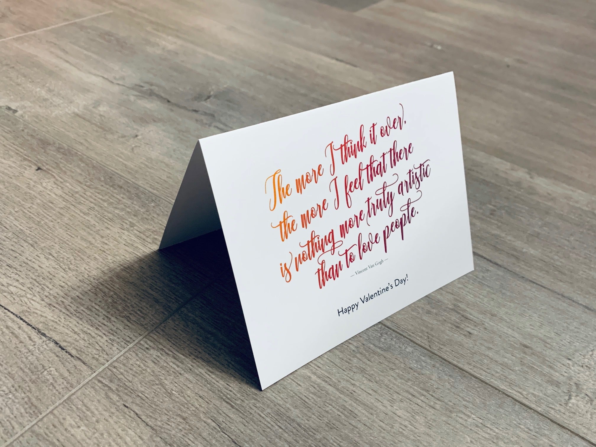 A folded white notecard is folded and propped up on a gray wooden background. The card says, "The more I think it over, the more I feel that there is nothing more truly artistic than to love people. Happy Valentine's Day!" Historic Love collection by Stationare.