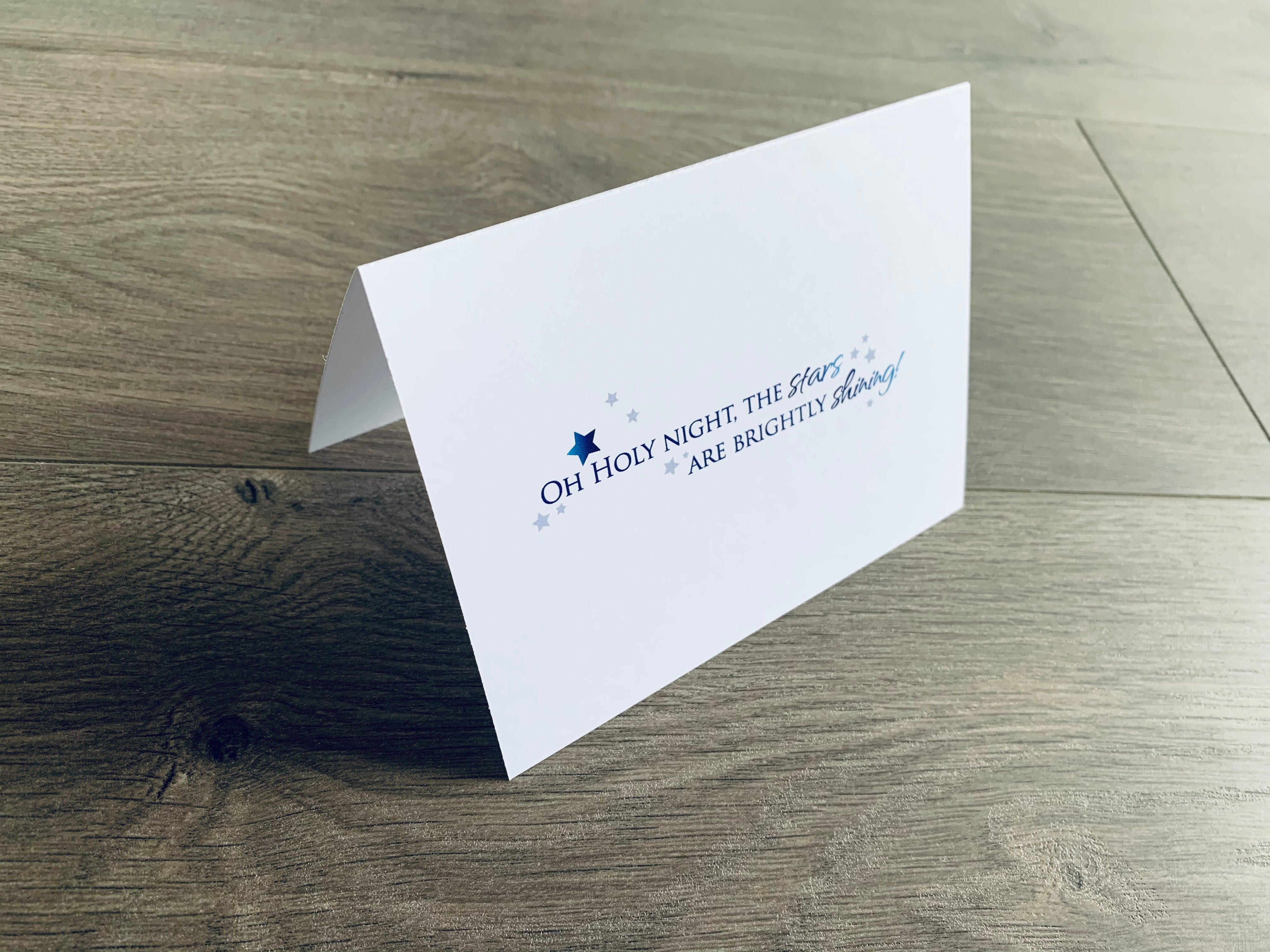 A white, folded notecard sits on a wooden floor. The card says, "Oh holy night, the stars are brightly shining" with small blue stars around the saying. Christmas Magic collection by Stationare.