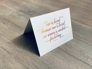 A folded white notecard is folded and propped up on a gray wooden background. The card says, "One is loved because one is loved. No reason is needed for loving." Historic Love collection by Stationare.