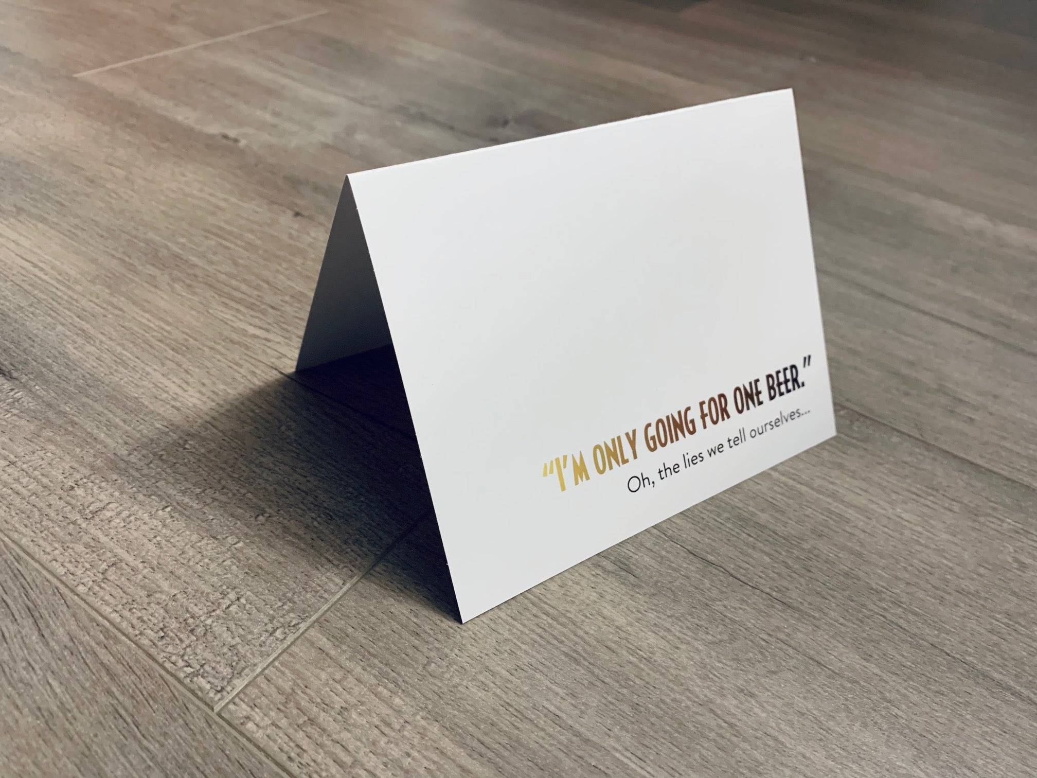 A white card is propped on a gray wood floor. The card reads, "I'm only going for one beer. Oh, the lies we tell ourselves..." Beer Lovers collection by Stationare.