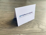 A white folded notecard is propped up on a wooden floor. The card reads "opportunities are everywhere" in a handwritten-style font that fades from blue to purple. Below it are capitalized letters that read "look for them. Create them. Embrace them." From Stationare's Inspire Collection.