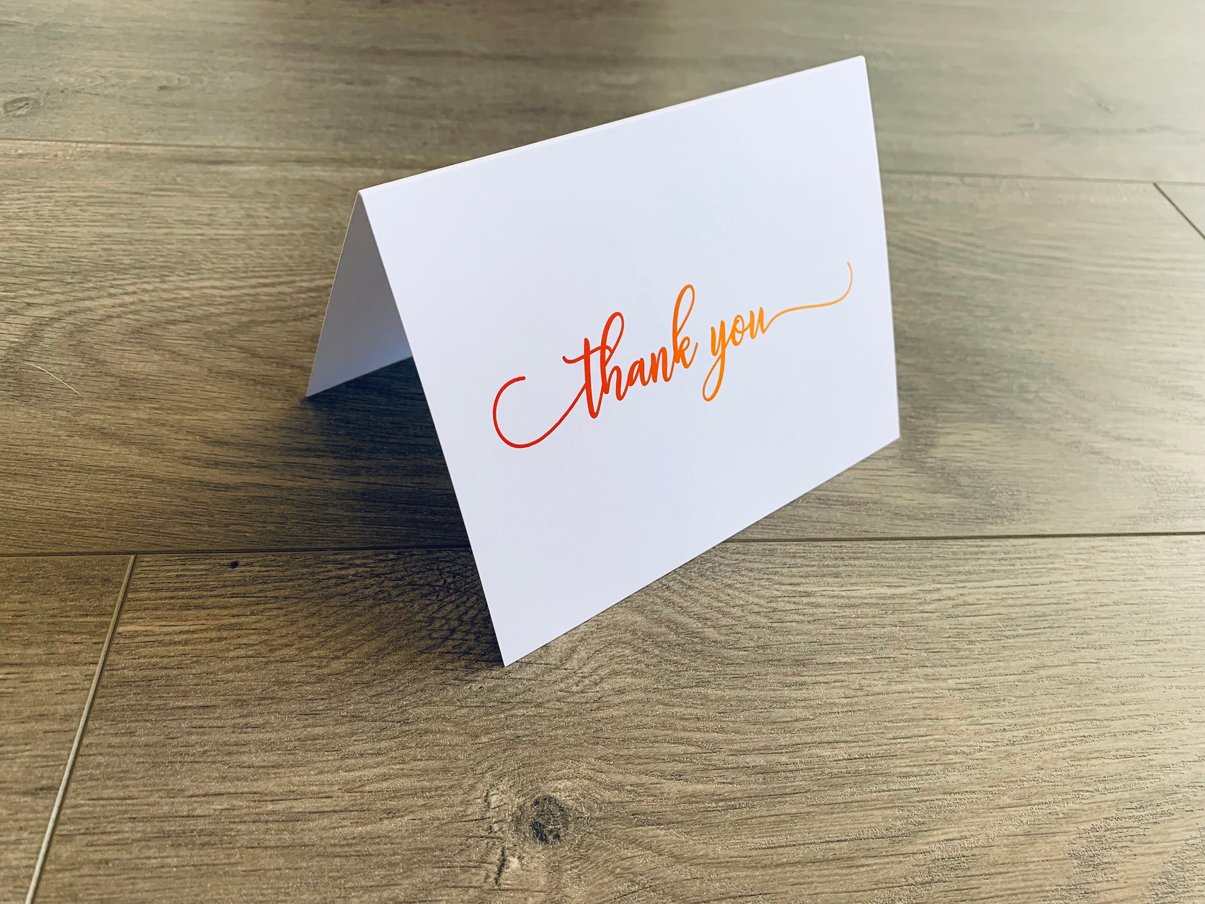 A white notecard with a script font that reads "thank you" on a wooden floor. The font fades from dark to light orange.