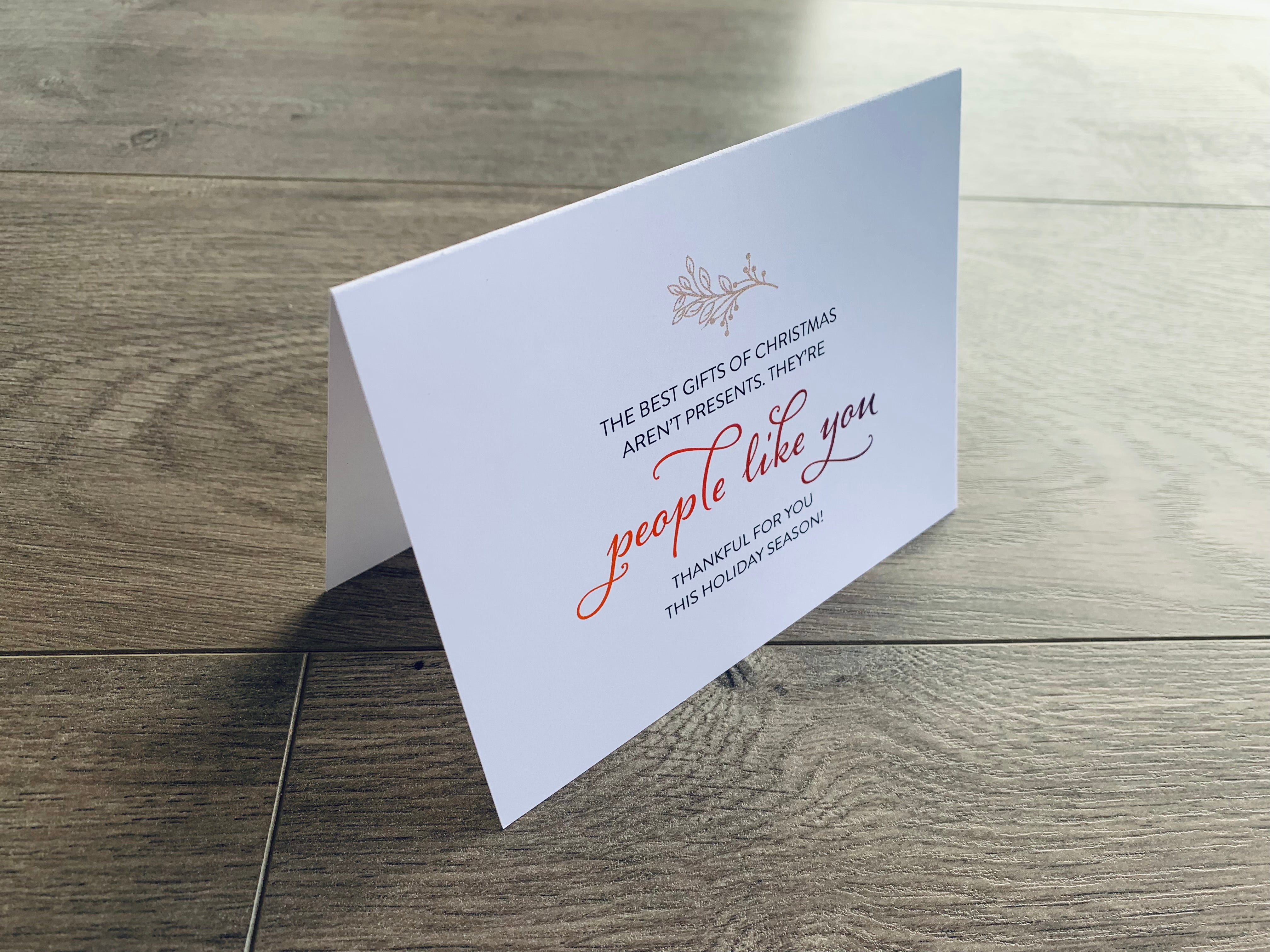 A white folded notecard sits on a wooden floor. On the front of the card, it reads, "The best gifts of Christmas aren't presents. They're people like you. Thankful for you this holiday season!" Merry Thanks Collection by Stationare.