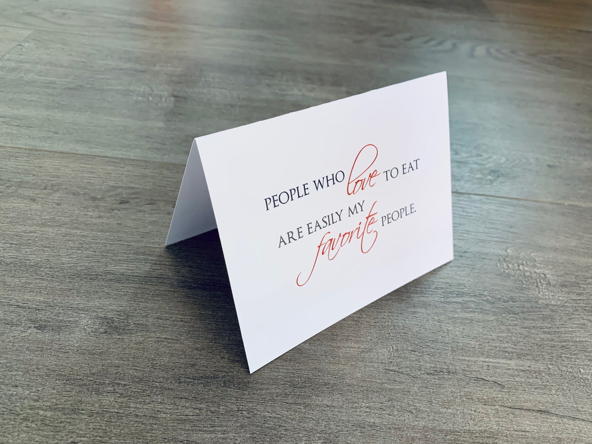 A white, folded notecard sits on a wooden floor. The card says, "People who love to eat are easily my favorite people." The Foodie collection by Stationare.
