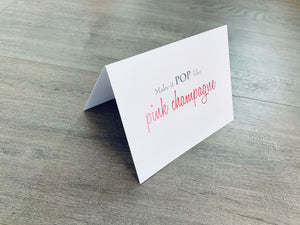 A white notecard is on a gray wooden floor. The card says, "Make it POP like pink champagne." Champagne Lovers collection by Stationare.