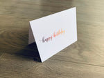 A single white notecard is propped up on a wooden floor. The card reads, "happy birthday" in a fancy script font in purple to orange ombre. Happy Birthday Collection by Stationare.