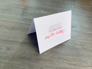 A white notecard is on a gray wooden floor. The card says, "Why have a party when you can have a celebration? pop the bubbly." Champagne Lovers collection by Stationare.