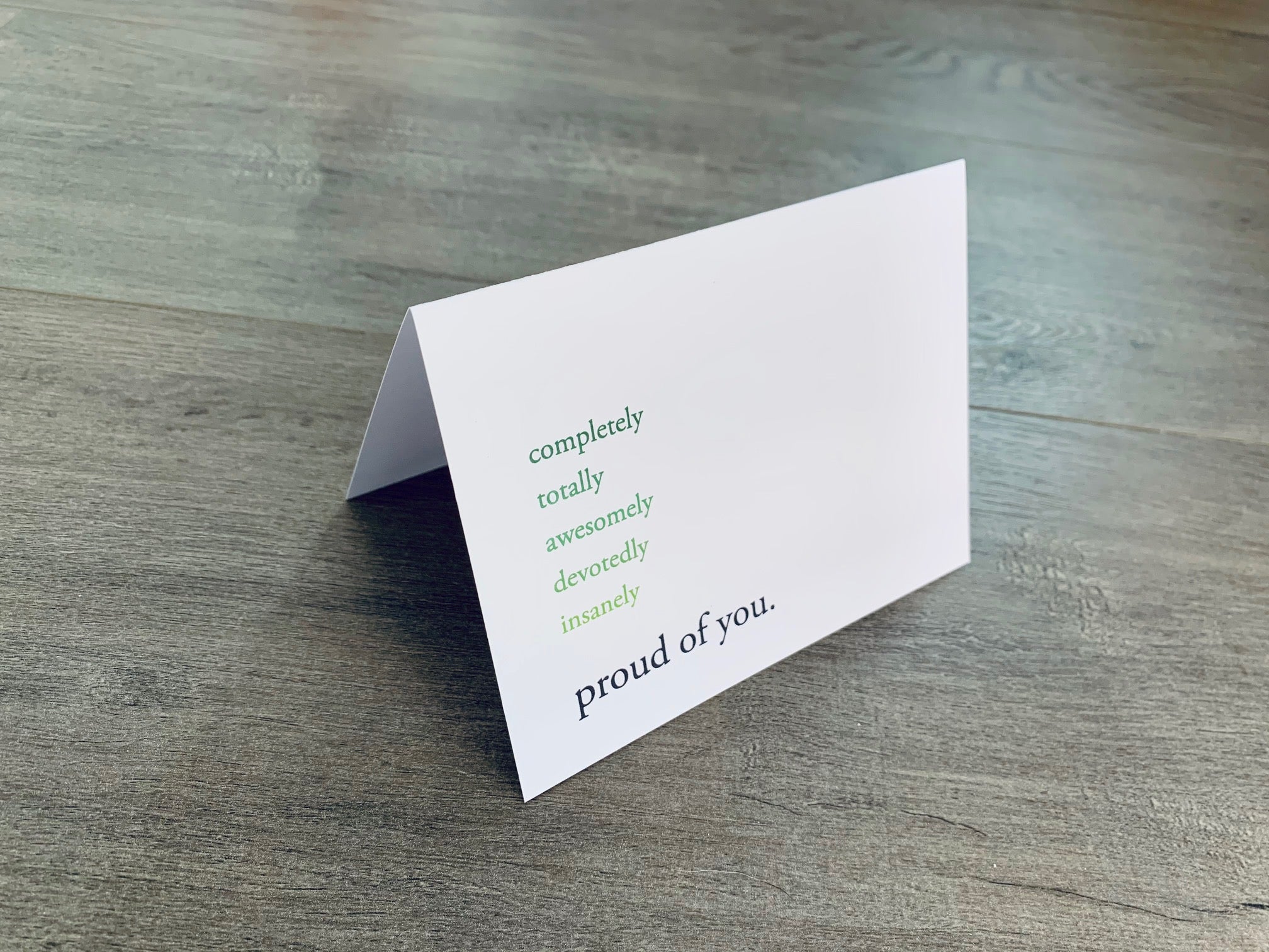 A white, folded notecard sits on a wooden floor. The card says, "completely. totally. awesomely. devotedly. insanely. proud of you." Congrats collection by Stationare.