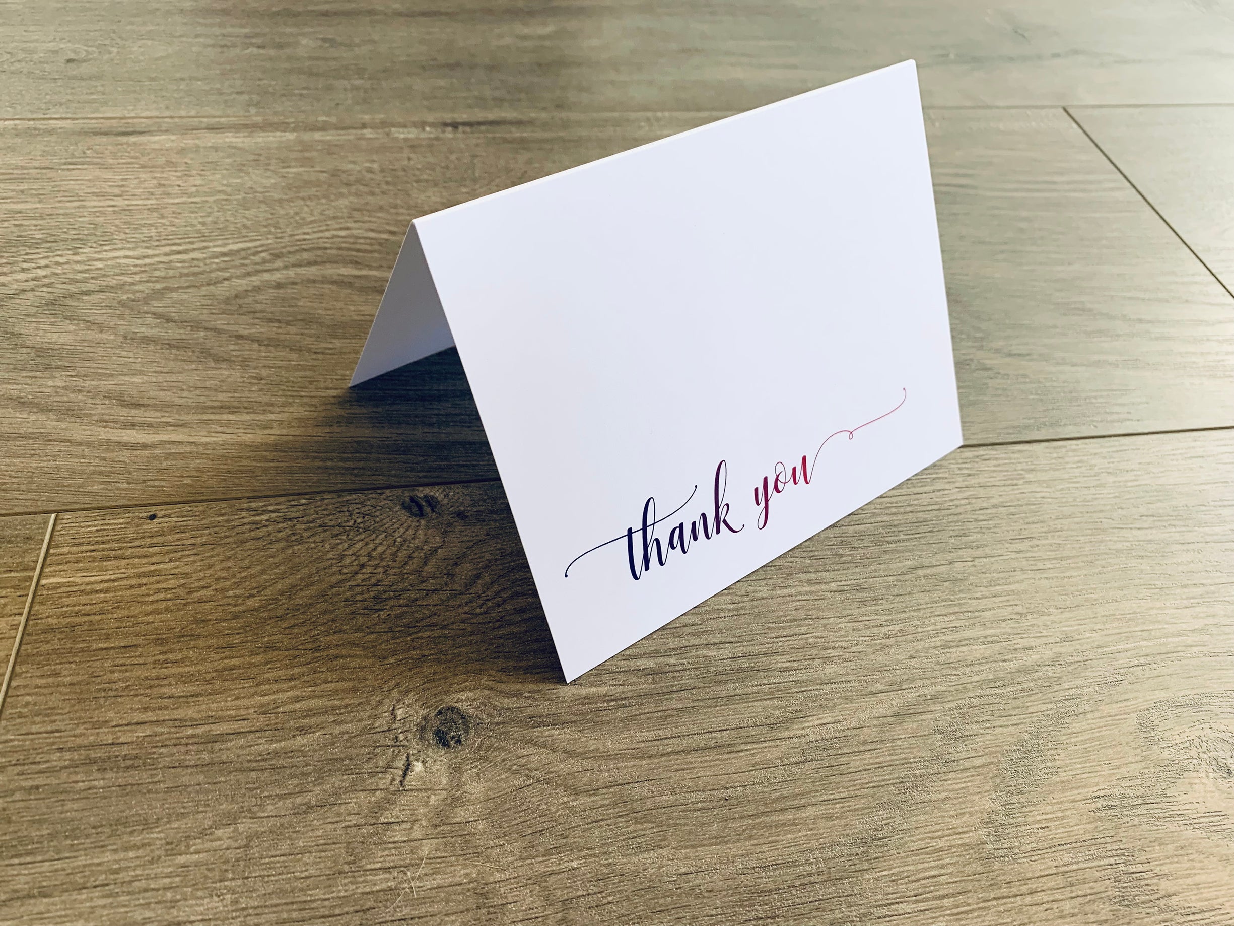 A white notecard with a script font is propped up on a wooden floor. The card reads "thank you." The font fades from purple to pink. 