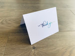A white notecard with a script font that reads "thank you" is folded and propped up on a wooden floor. The font fades from navy to lighter blues to lime green.