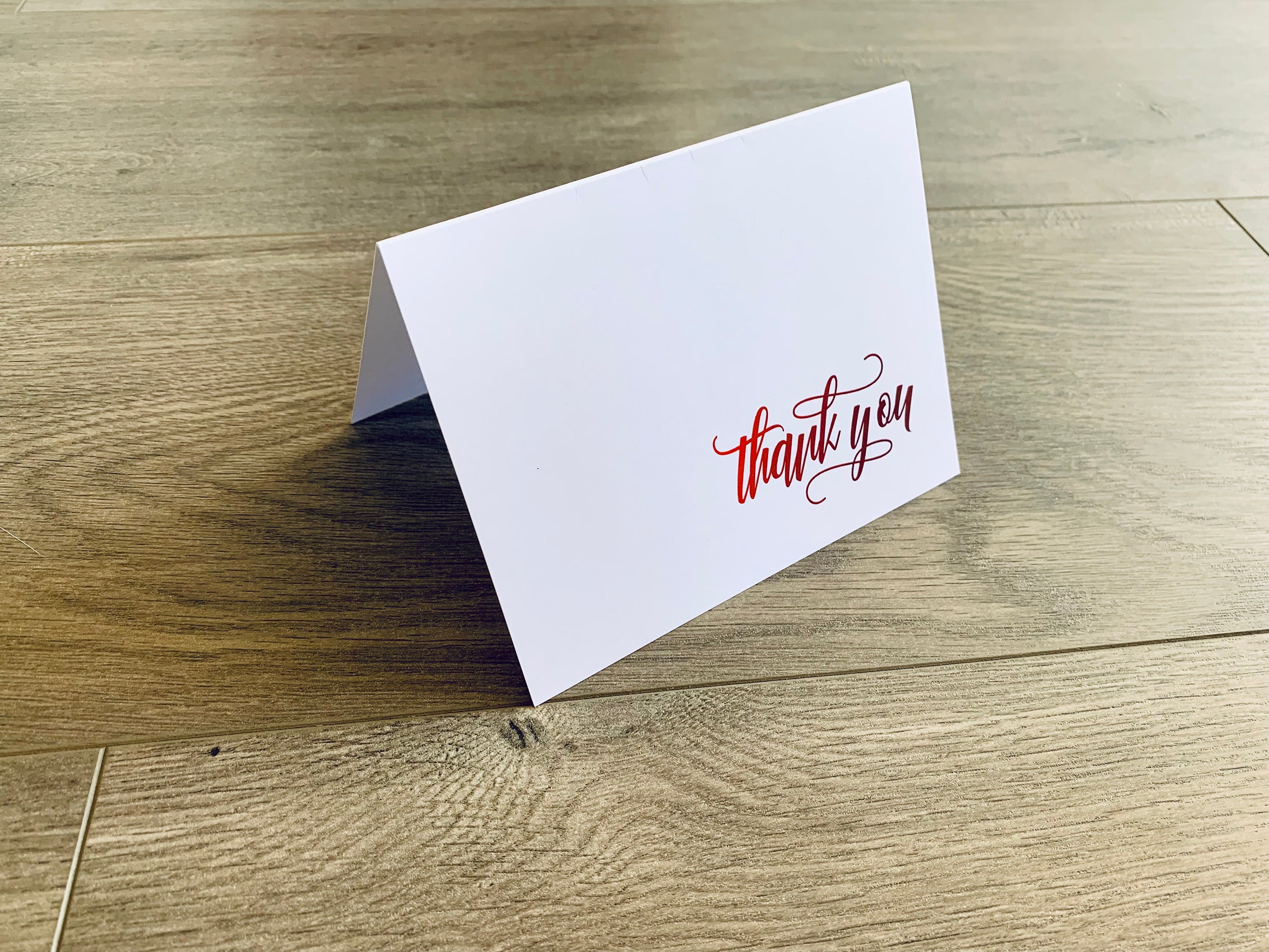 A white notecard with a script font that reads "thank you" sits on a wooden floor. The font fades from bright red to dark burgundy.