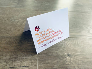 A folded white notecard is folded and propped up on a gray wooden background. The card says, "Roses are red, violets are blue. Thanks for feeding me... and picking up my poo. Happy Valentine's Day!" Valentines from the Pup collection by Stationare.