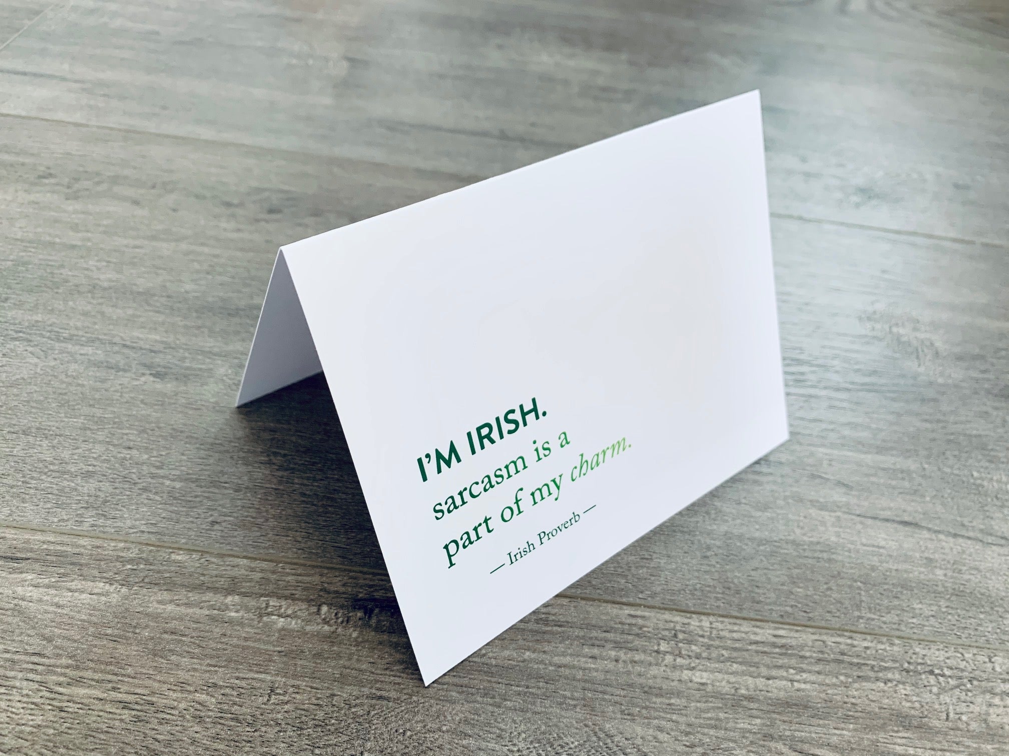 A white folded notecard is propped up on a wooden floor. The card has the Irish proverb, "I'm Irish. Sarcasm is a part of my charm." From the Irish Laughs Collection by Stationare.