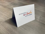 A folded white notecard is propped up on a wooden floor. The card reads, "See the world as a traveler not as a tourist." Stationare's Travel Lovers collection.