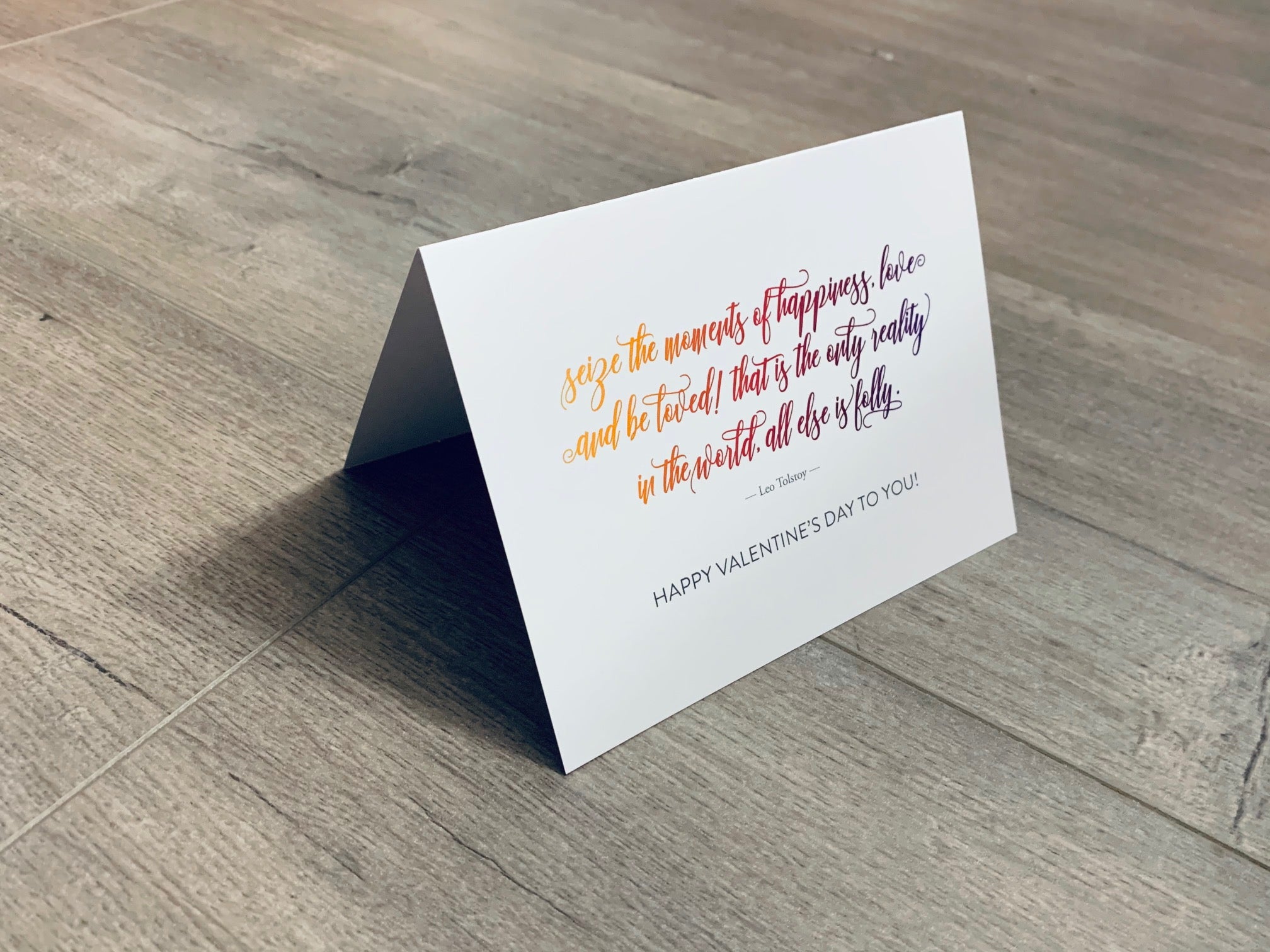 A folded white notecard is folded and propped up on a gray wooden background. The card says, "Seize the moments of happiness, love and be loved! That is the only reality in the world, all else is folly. Happy Valentine's Day to you!" Historic Love collection by Stationare.