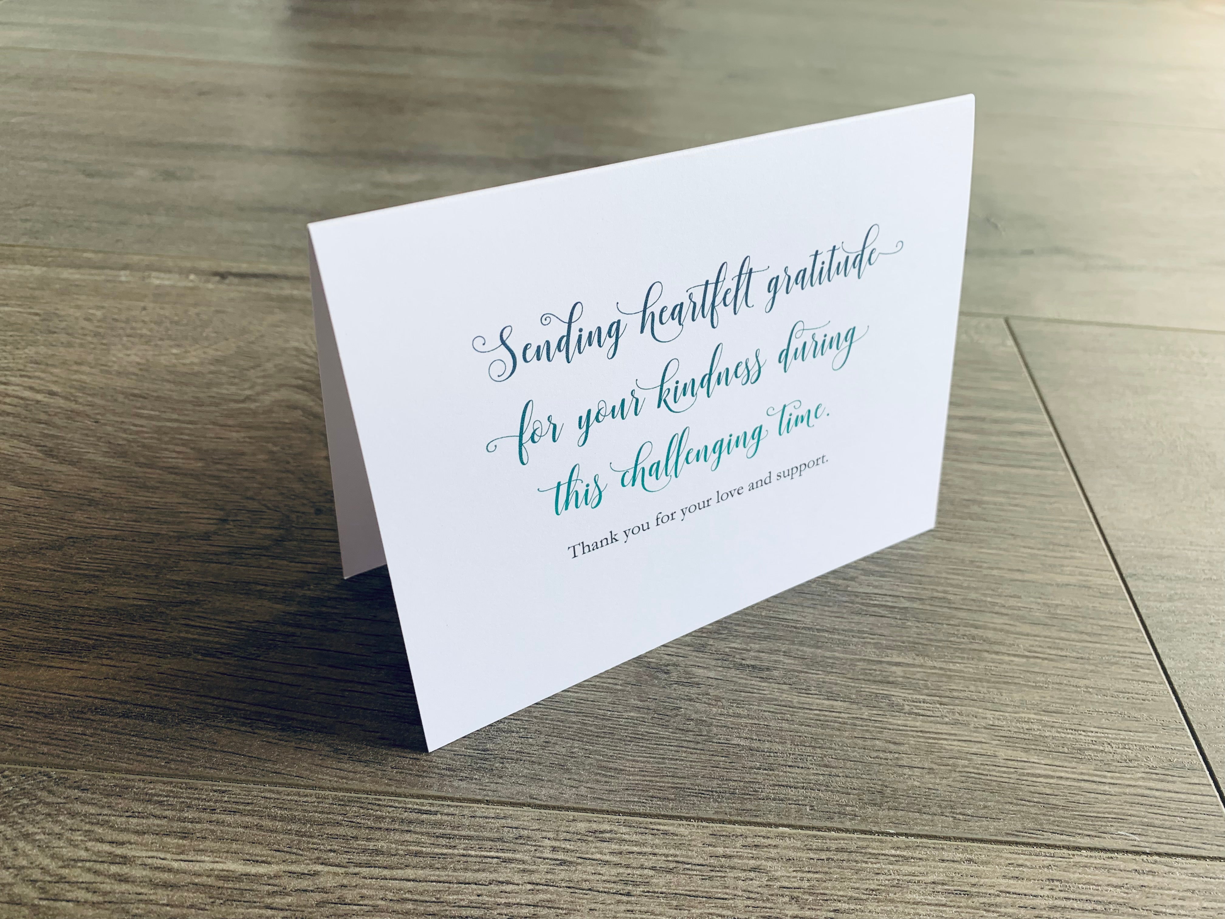 A folded white notecard is propped up on a wooden floor. The card reads, "Sending heartfelt gratitude for your kindness during this challenging time. Thank you for your love and support." Stationare's Surviving Hard Times collection.
