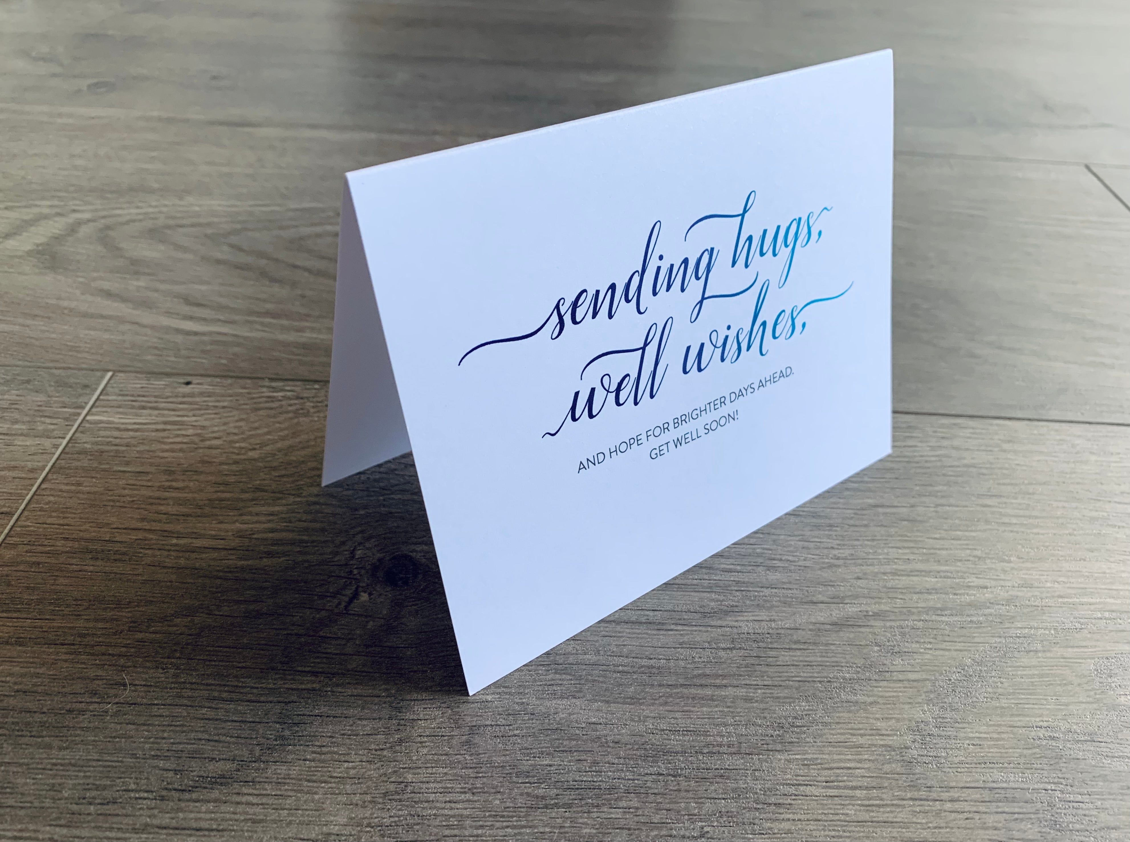 A white notecard that reads, "Sending hugs, well wishes and hope for brighter  days ahead. Get well soon!" is folded and propped up on a gray wood floor. From Stationare's Get Well Soon collection